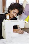 Young hispanic female dressmaker using sewing machine and smiling at camera. Vertical shape, side view, waist up