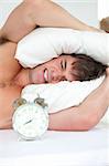 stressed man woken-up by his alarm clock putting his head under the pillow in the morning