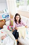 Joyful woman sitting on the sofa with bags reading a card while her baby is sleeping in his cradle