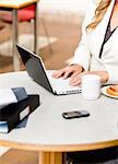Close-up of a busy businesswoman using her laptop with cellphone, mug and food on the table in her office