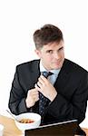 Elegant businessman having breakfast and touching his tie in front of his laptop against a white background