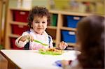 Caucasian and hispanic female preschoolers eating pasta and smiling. Horizontal shape, waist up, focus on background