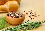 Black, white, green and red pepper corns in a ceramic bowl with other herbs (rosemary, dill, thyme) in the foreground and a mortar and pestle in the background (Selective Focus, Focus on the front of the bowl and the pepper corns in the bowl)