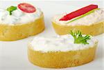 Baguette bread with cream cheese spread decorated with tomato, red pepper, parsley and green onion on white plate (selective focus)
