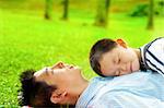 Boy lying on father. Father lying on green grass outside.