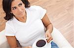 A beautiful young Hispanic Latina woman on her sofa drinking tea or coffee from a white cup and saucer