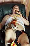 Photo of a fat couch potato eating a huge hamburger and watching television.  Harsh lighting from the television illuminates the dark room.