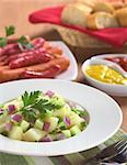 Fresh potato salad with cucumber, red and green onion made with an oil dressing with barbecued sausages, mustard, ketchup and baguette in the back (Selective Focus, Focus on the front of the salad and the parsley leaf)