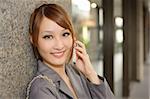 Attractive young business woman holding cellphone against wall and looking at you, half length closeup portrait outside of modern buildings.