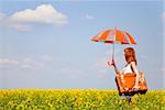 Redhead enchantress with umbrella and suitcase at spring rapeseed field.