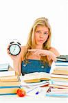 Worried teen girl sitting at table with books and pointing finger on alarm clock
