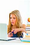 Pensive young girl sitting at table with books and looking on laptop