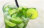 The cocktail Mojito with rum, limes, mint and ice cubes photographed on sand (Selective Focus, Focus on the mint leaf and the upper part of the lime slice on the rim)