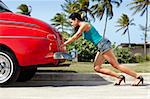 young hispanic woman pushing broken down red convertible vintage car. Horizontal shape, full length, side view