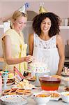 Two women at party putting candles in cake smiling