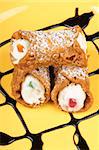 Close-up of original mini sicilian cannoli over a yellow plate. These tube-shaped pastries are made of fried shells filled with a sweet mixture of ricotta cheese, chocolate, candied fruit and sprinkled with icing sugar.