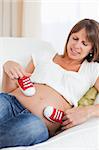 Gorgeous pregnant female playing with red baby shoes while lying on a sofa in her living room