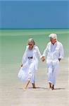 Happy senior man and woman couple walking, smiling and holding hands on a deserted tropical beach with bright clear blue sky