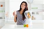 Beautiful woman using a blender while holding a drink in the kitchen