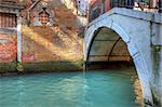 Small canal flow along old brick walls under vintage bridge in Venice, Italy.