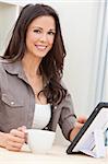 Beautiful, smiling, young brunette woman at home at a table using her tablet computer or iPad drinking a mug of tea or coffee