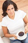 A beautiful young Latina Hispanic woman or girl with an enigmatic smile drinking tea or coffee from a white cup at home on her sofa