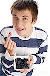 A boy holding a single blueberry in his fingers and smiling, while holding a small bowl filled with the delicious blueberries.