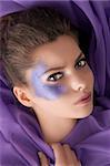 beauty shot of a young girl with purple glitter make up on her eyes laying on purple satin