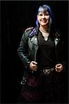 Portrait of smiling Caucasian woman with blue hair and black leather jacket standing against black background.