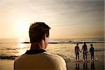 Caucasian mid-adult man standing and watching mid-adult woman with children on beach.