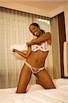 Young African American woman kneeling on bed in lingerie holding pillow and smiling at viewer.