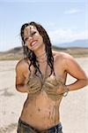 Topless Caucasian mid-adult woman holding breasts covered in mud in desert smiling and looking at viewer.