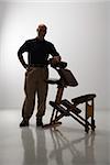 Silhouette of Caucasian middle-aged male massage therapist standing with elbow on massage chair.