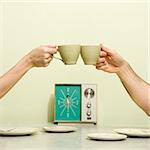 Caucasian male and female hands toasting with coffee cups across retro kitchen table setting.