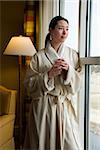 Taiwanese mid adult woman in bathrobe looking out window holding coffee cup.