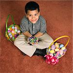 Hispanic boy sitting on floor with two Easter baskets holding chocolate candy eggs in his hands looking up at viewer smiling.