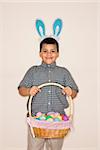 Hispanic boy wearing bunny ears holding Easter basket smiling and looking at viewer.