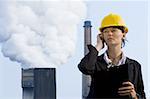 A beautiful dark haired woman wearing a hard hat and talking on her mobile phone while standing in front of a factory pumping out pollution from its chimneys