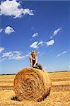 Woman sitting on a hay bale in summer stubble field under a bright vivid sky