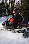 Young man sitting on snowmobile in snow with helmet on lap smiling.