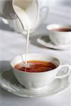 Pouring milk from porcelain milk jug into cup filled with tea - white tablecloth background.