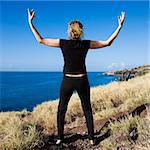 Back view of Caucasian mid-adult woman standing on Maui, Hawaii coast with arms raised above head.