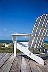 Empty white adirondack chair on wooden deck facing beach.