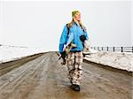 Young woman in winter clothes walking down muddy dirt road holding snowboard and boots smiling.