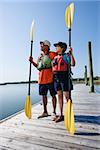 African American middle-aged couple standing on boat dock holding paddles wearing life preservers.
