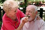 A senior woman caring for her husband and feeding him a grape.