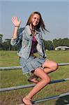 a beautiful girl sitting on a fence by a farm, waving.