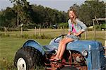 A beautiful blond girl sitting on a tractor in the middle of a farm.