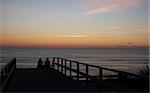 Photograph of a wooden walkway leading down to the beach with a romantic couple sitting at the end.