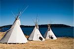 Three teepees are lined up in a row in front of water. These teepees are used to provide shelter at a summer camp. Teepees were one form of traditional shelter used by native americans.
